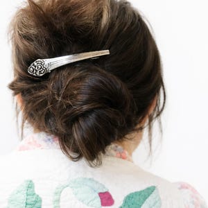 May include: Silver-toned hair barrette, crafted from a spoon, is worn in a bun. The barrette features a decorative floral design on the spoon's handle. The person is wearing a white jacket with floral embroidery.