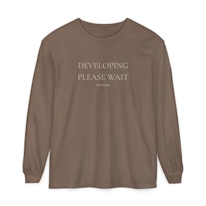 May include: A long-sleeved, brown t-shirt with the text "DEVELOPING PLEASE WAIT" and "8-12 hours" printed in the center. The shirt has a crew neck and a relaxed fit, suitable for casual wear.