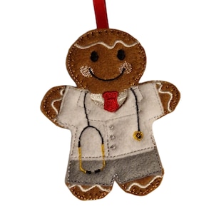 May include: A felt gingerbread man decoration wearing a white lab coat, grey trousers, and a red tie. The decoration has a stethoscope and a red ribbon for hanging. The gingerbread man has a smiling face.