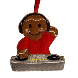Peut inclure: Ornement de Noël en feutre en forme de bonhomme en pain d'épices DJ. Le bonhomme en pain d'épices porte un casque, une chemise rouge et se tient derrière une table de mixage grise. Un ruban rouge est attaché pour l'accrocher.