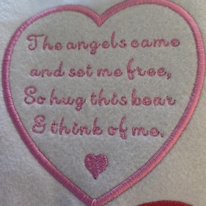 Könnte beinhalten: Ein weißer Stoffaufnäher in Herzform mit einem rosa gestickten Rand. Der Text lautet: The angels came and set me free, So hug this bear & think of me. Unter dem Text befindet sich ein kleines, ausgefülltes, rosa gesticktes Herz auf weißem Grund.