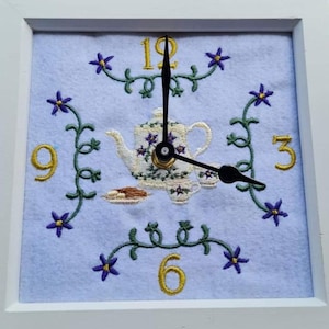 Può includere: Orologio da parete quadrato con cornice bianca e sfondo azzurro. Il quadrante presenta un disegno ricamato di una teiera, tazzine e decorazioni floreali. I numeri 3, 6, 9 e 12 sono dorati. Le lancette dell'orologio sono nere.