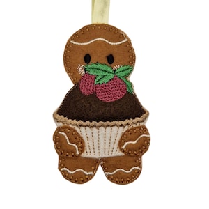 May include: A brown felt gingerbread man ornament with a cupcake body. The gingerbread man has a stitched white icing design, black eyes, and a green and red embroidered berry garnish. A yellow ribbon is attached for hanging.