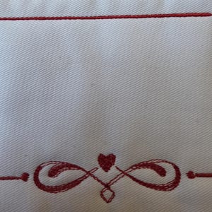 May include: White fabric featuring a red embroidered border and decorative elements. The design includes a rectangular frame and a heart with infinity symbols. The embroidery is detailed, and the fabric has a subtle texture.