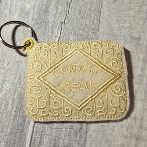 May include: A cream-colored, embroidered keychain shaped like a custard cream biscuit. The biscuit has a diamond-shaped center with the words "CUSTARD CREAM" embroidered in a matching cream color. The edges feature an ornate, swirling design, and a silver keyring is attached.
