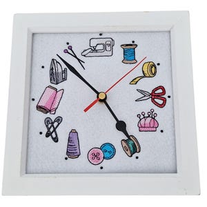 Op de afbeelding: Een vierkante klok met een wit frame en een witte wijzerplaat met geborduurde iconen met naaigerei. De wijzers zijn zwart met een rode secondewijzer. De iconen omvatten een naaimachine, schaar, garenklosjes en een speldenkussen.
