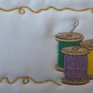 Può includere: Ricamo su tessuto bianco con tre rocchetti di filo verde, viola e giallo. Un bordo decorativo dorato circonda uno spazio vuoto, suggerendo un oggetto personalizzato o un progetto artigianale.