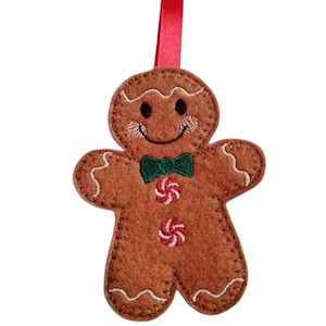 Può includere: Un ornamento natalizio a forma di omino di pan di zenzero in feltro marrone con un nastro rosso per appenderlo. L'ornamento ha una faccia sorridente, un papillon verde e bottoni di caramelle rosse e bianche. I bordi sono cuciti con filo bianco.