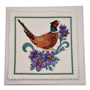 Op de afbeelding: Geborduurde wenskaart met een fazant met oranje, bruine en zwarte veren, een rode kop en een groene nek. De fazant zit op een tak met paarse bloemen en groene bladeren. De kaart heeft een witte achtergrond met een blauwgroene rand.