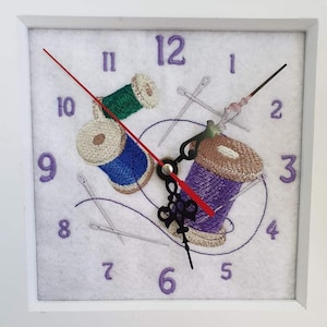 Può includere: Orologio quadrato bianco con design ricamato con rocchetti di filo blu, verde e viola, insieme ad aghi. Il quadrante dell'orologio ha numeri viola e lancette nere con una lancetta dei secondi rossa. Un oggetto unico, fatto a mano.