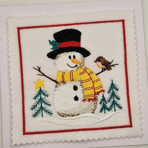 May include: Embroidered snowman design on a white square card. The snowman wears a black top hat, yellow scarf, and has a bird perched on its arm. Includes two green Christmas trees and a yellow star. The card is framed with a red border.