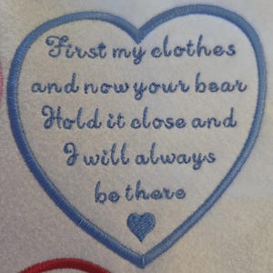 Könnte beinhalten: Ein weißer Stoffaufnäher in Herzform mit blauem Stickrand. Der Text lautet: First my clothes and now your bear Hold it close and I will always be there. Ein kleines blaues Herz ist unter dem Text mit blauem Garn eingestickt.