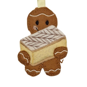 May include: A brown felt gingerbread man decoration holding a light yellow cake with a white and brown embroidered design. The ornament has a cream-coloured ribbon for hanging, ideal for festive decor.