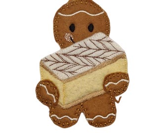 Op de afbeelding: Een bruin vilten peperkoekmannetje ornament met een lichtgele cake met een wit en bruin geborduurd ontwerp. Het ornament heeft een crèmekleurig lint om op te hangen, ideaal voor feestelijke decoratie.