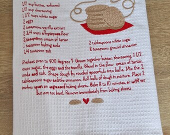 May include: White waffle-weave kitchen towel with embroidered recipe for Snickerdoodles cookies in red thread. The recipe includes ingredients and instructions. A stack of cookies is embroidered on the towel.