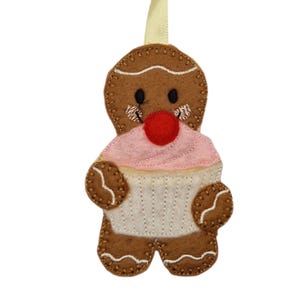 May include: A felt gingerbread man Christmas decoration. The brown gingerbread man holds a pink cupcake with a red cherry. The ornament has a cream ribbon for hanging. The stitching is visible.
