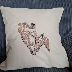 May include: A cream-coloured square cushion featuring an embroidered design of two giraffes. The giraffes are in shades of brown, with detailed patterns on their coats. The cushion is on a blue corduroy surface.