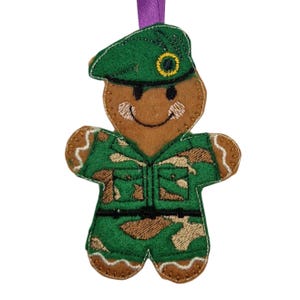 May include: A gingerbread man ornament wearing a green military uniform and beret. The ornament has a smiling face and is decorated with brown, green, and tan felt. A purple ribbon is attached for hanging.
