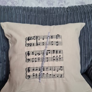 Puede incluir: Cojín color crema con notas musicales bordadas y una flauta. Las notas musicales negras están dispuestas en tres filas, con una flauta plateada superpuesta a las notas. El cojín está sobre un sofá de pana azul.