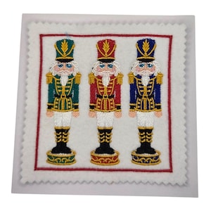 Könnte beinhalten: Gesticktes Quadrat mit drei Spielzeugsoldaten-Nussknackern in grünen, roten und blauen Uniformen. Jeder Nussknacker hat einen weißen Bart, einen hohen Hut und goldene Akzente. Das Design wird von einem roten Rand auf weißem Hintergrund umrahmt.