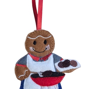 May include: A felt gingerbread man Christmas ornament with a red ribbon. The gingerbread man is wearing an apron and grilling food on a red barbecue. The ornament shows the gingerbread man holding a plate of food.