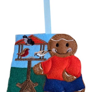 May include: A felt Christmas decoration depicting a gingerbread man in a red top and blue trousers. The ornament has a light blue ribbon. A scene with birds at a bird table is on the left side of the decoration.