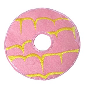 May include: A pink donut-shaped plush toy with yellow frosting details. The toy has a circular shape with a hole in the centre and yellow embroidered frosting dripping down the sides. The edges are finished with a stitched border.
