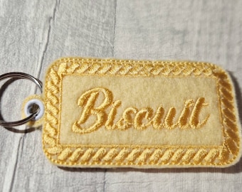 Op de afbeelding: Een gele rechthoekige sleutelhanger met het woord "Biscuit" in sierlijke letters geborduurd. De sleutelhanger heeft een decoratieve rand en een zilveren sleutelring.