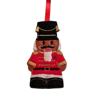 May include: A felt Christmas ornament in the shape of a nutcracker soldier. The ornament has a black hat, red jacket with gold trim, black trousers, and a white beard. A red ribbon is attached for hanging.