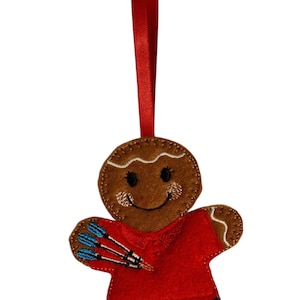 Könnte beinhalten: Ein Weihnachtsornament aus Filz, das einen Lebkuchenmann mit rotem Hemd, blauen Shorts und drei blau-weißen Darts darstellt. Das Ornament hat ein rotes Band zum Aufhängen und ist etwa 10 cm hoch.