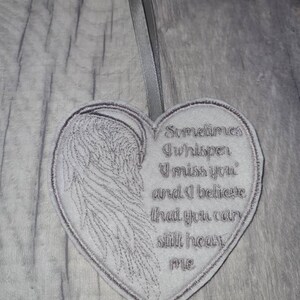 May include: A heart-shaped grey fabric ornament featuring an embroidered angel wing on the left side and the text 'Sometimes I whisper I miss you and I believe that you can still hear me' on the right, finished with a grey ribbon loop for hanging.