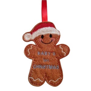 May include: A festive gingerbread man Christmas decoration with a red and white Santa hat and a red hanging ribbon. The ornament features the embroidered text "BABY'S 1st CHRISTMAS".