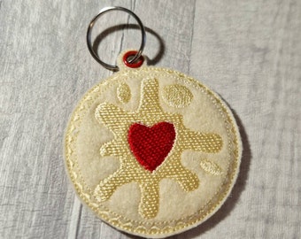 Op de afbeelding: Een crème-kleurige, geborduurde sleutelhanger met een rood hart in het midden. De sleutelhanger heeft een cirkelvorm met een decoratief ontwerp rond het hart. Een zilveren sleutelring is aan de bovenkant bevestigd met een rode ring.