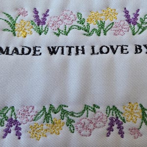 May include: White fabric featuring embroidered floral borders in yellow, purple, pink, and green. The text "MADE WITH LOVE BY" is embroidered in black, positioned between the floral designs. The fabric has a textured appearance.