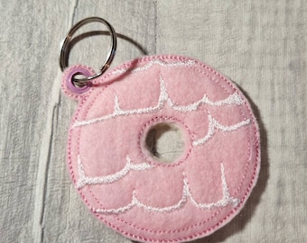 Op de afbeelding: Een roze vilten donut sleutelhanger met witte geborduurde glazuur details en een zilveren sleutelring. De sleutelhanger is rond met een gat in het midden en een geschulpte rand. De donut meet ongeveer 7.6 cm in diameter.
