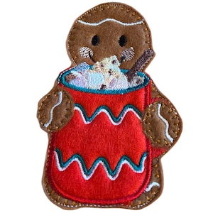May include: A felt gingerbread man with a red apron, holding a mug of hot chocolate. The gingerbread man is brown with white icing details. The mug is red with a teal and white wavy design.