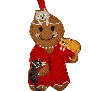 May include: A festive gingerbread-shaped Christmas decoration featuring a smiling face and three embroidered cats. The ornament has a red dress and a red ribbon for hanging. The cats are black, orange, and white.