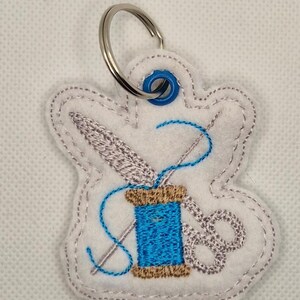 May include: White felt keychain with a sewing theme. The design features a blue spool of thread, a needle, and scissors, all embroidered in detail. A silver key ring is attached at the top. The keychain is a craft item.