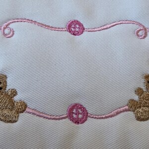May include: Embroidered design on white fabric, featuring two brown teddy bears and a pink frame. The frame has decorative swirls and circular accents. The teddy bears have pink ribbons. The design is suitable for baby items.