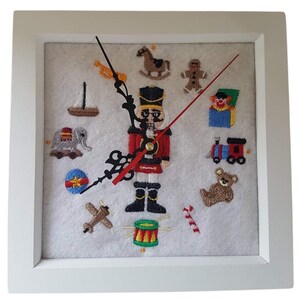 Könnte beinhalten: Quadratische, weiß gerahmte Uhr mit einem Filzhintergrund mit gestickten Spielzeugmotiven. Das Zifferblatt der Uhr zeigt einen Nussknacker, eine Spielzeugeisenbahn, einen Teddybären und andere farbenfrohe Stickereien. Die Zeiger der Uhr sind schwarz und rot.
