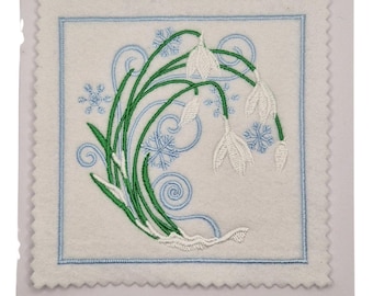 May include: Embroidered square card featuring snowdrop flowers, green stems, and blue snowflakes. The design is framed by a light blue border and set against a white background. The card has a scalloped edge.