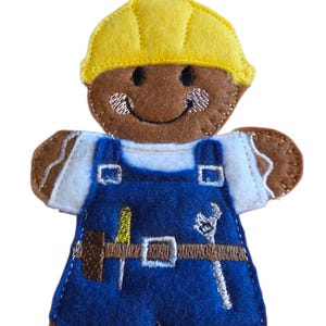 Peut inclure: Ornement en feutre en forme de bonhomme en pain d'épices représentant un ouvrier du bâtiment. Il porte un casque jaune, une salopette bleue et des outils. Le bonhomme en pain d'épices a un visage souriant et est bordé de coutures marron.