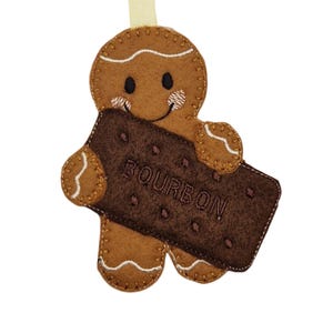 Puede incluir: Adorno de hombre de jengibre de fieltro que sostiene una galleta de fieltro marrón oscuro con la palabra "BOURBON" cosida. El hombre de jengibre tiene una cara sonriente, detalles de glaseado blanco y una cinta beige para colgar.