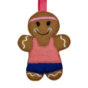 May include: A gingerbread person decoration with a pink headband, vest, and blue shorts. The ornament has a pink ribbon for hanging and is decorated with white stitching.