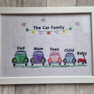 以下が含まれることがあります： 白いフレームに入った刺繍アート。 「The Car Family」の文字が上にあり、家族の各メンバーを表す緑、紫、ピンク、青の車が描かれています。赤ちゃんを表す赤いワゴンがあります。