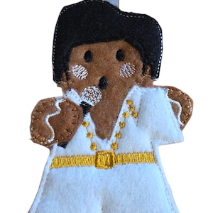 Pode incluir: Um enfeite de feltro em forma de cantor com um macacão branco com detalhes dourados e penteado preto. O enfeite tem uma fita azul clara para pendurar e um microfone prateado. A pele do cantor é castanha.