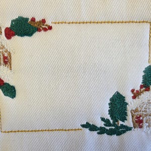 Op de afbeelding: Een wit stoffen oppervlak met een rechthoekige, goudkleurig geborduurde rand. Het ontwerp bevat twee geborduurde huisjes met besneeuwde daken, groene dennenbomen en rode bessen in de linker bovenhoek en de rechter benedenhoek.