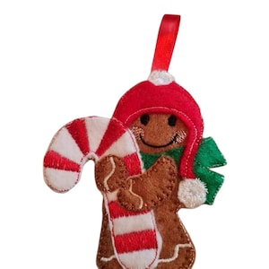 Könnte beinhalten: Ein Weihnachtsornament aus Filz, das einen Lebkuchenmann darstellt. Der braune Lebkuchenmann trägt eine rote Mütze und einen grünen Schal und hält eine rot-weiße Zuckerstange. Ein rotes Band ist zum Aufhängen befestigt.