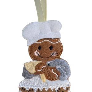 May include: A felt gingerbread man ornament wearing a chef's hat and apron, holding a spatula. The ornament is brown, white, and grey, with a light yellow ribbon for hanging. The gingerbread man is standing on a cake.