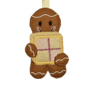 Könnte beinhalten: Ein brauner Lebkuchenmann-Ornament aus Filz, der ein quadratisches Geschenk hält. Das Ornament hat weiße Zuckergussdetails, schwarze Augen und gestickte Details. Ein gelbes Band ist zum Aufhängen befestigt. Das Geschenk hat einen gelben Rand und ein rosa-gelbes Karomuster.